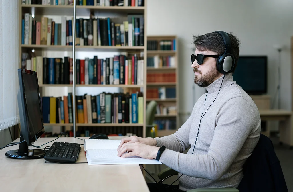 Homem cego, usando fones de ouvido, digita em um teclado ao lado de um livro em braille, em frente a um computador em uma biblioteca